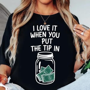 Puede incluir: Camiseta negra con la frase blanca "I LOVE IT WHEN YOU PUT THE TIP IN". Un gráfico de un tarro con dinero está debajo del texto. La camiseta es de estilo casual.
