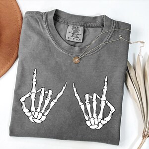 a gray t-shirt with a graphic design of two skeletal hands making the "rock on" sign, accompanied by a necklace and a hat.