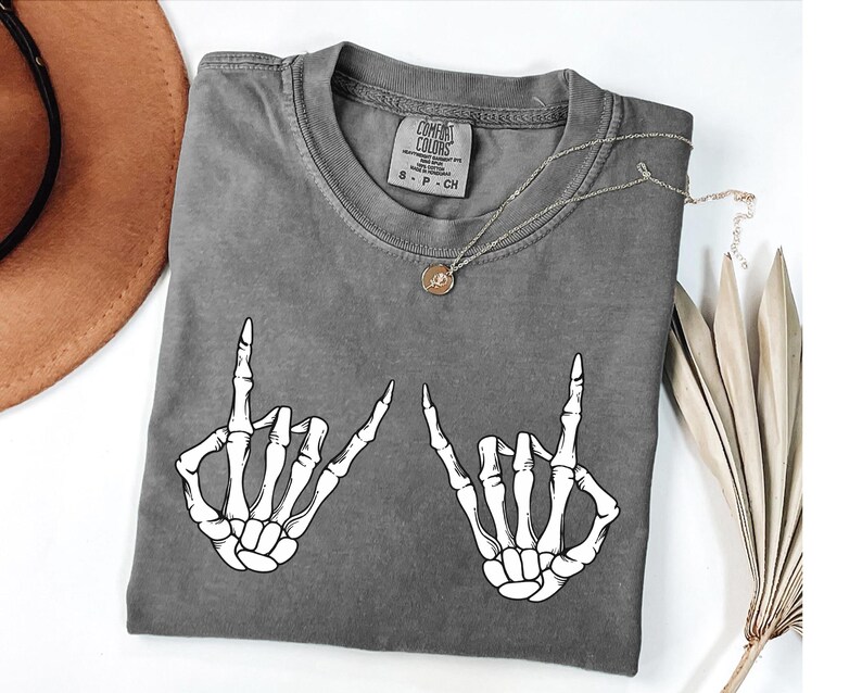 a gray t-shirt with a graphic design of two skeletal hands making the "rock on" sign, accompanied by a necklace and a hat.