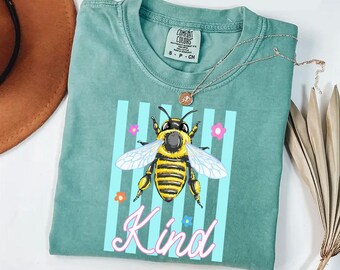 May include: A sage green short-sleeve t-shirt featuring a graphic of a yellow and black bee centred over light blue vertical stripes. Small pink, orange, and blue flowers surround the bee, with the word Kind written in pink script below.