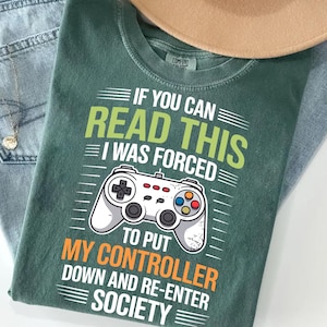 May include: Green t-shirt with the text "IF YOU CAN READ THIS I WAS FORCED TO PUT MY CONTROLLER DOWN AND RE-ENTER SOCIETY" and a graphic of a video game controller. The shirt is folded on top of a pair of blue jeans.
