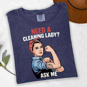 May include: Navy blue t-shirt with the text "Need a Cleaning Lady? Ask Me" and a graphic of a woman flexing her arm. The woman is wearing a red polka dot bandana and a blue shirt. The shirt is folded.