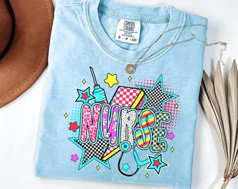 May include: A light blue t-shirt featuring the word NURSE in colourful, patterned block letters. The graphic includes a syringe, a stethoscope, and various star shapes in bright pink, yellow, and teal with checkered and leopard print details.