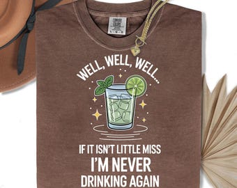 May include: Brown t-shirt with the text "WELL, WELL, WELL... IF IT ISN'T LITTLE MISS I'M NEVER DRINKING AGAIN." The graphic features a cocktail with ice, a lime slice, and mint. A gold necklace and a brown hat are also visible.