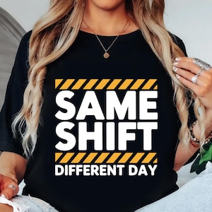 May include: Black t-shirt with the text "SAME SHIFT DIFFERENT DAY" in white block letters, framed by yellow and black diagonal stripes. The shirt is a casual style.