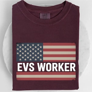 May include: A maroon t-shirt featuring an American flag design with the text "EVS WORKER" in white. The flag design includes red and white stripes and a blue field with white stars. The shirt is made of a soft material.
