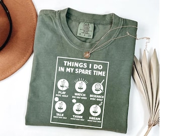 May include: An olive green t-shirt with the text "THINGS I DO IN MY SPARE TIME" and disc golf illustrations. The shirt has a Comfort Colors brand tag. A gold necklace is draped across the shirt. The shirt is a small size.