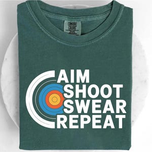 May include: A dark teal t-shirt with the words "AIM SHOOT SWEAR REPEAT" in white, along with a target graphic in blue, orange, and yellow. The shirt is made by Comfort Colors.