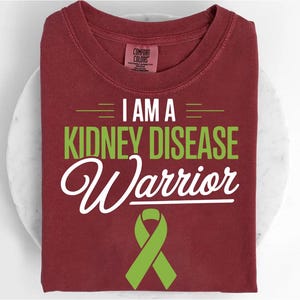 May include: A maroon t-shirt with the words "I AM A KIDNEY DISEASE Warrior" in green and white. A green ribbon is at the bottom. The shirt is folded and rests on a white surface.