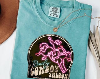 May include: A teal short-sleeved t-shirt featuring a graphic of a neon pink cowboy riding a bucking horse inside a circular frame. The text reads Rowdy COWBOY SALOON in a distressed font. The shirt has a Comfort Colors label at the inner neckline.