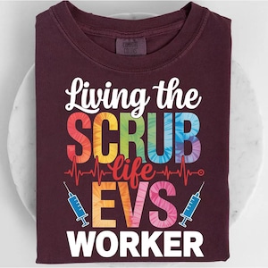 May include: A maroon t-shirt with the text "Living the SCRUB life EVS WORKER" in white and rainbow colors. The design includes a heartbeat line and two blue syringes. The shirt is folded on a white surface.