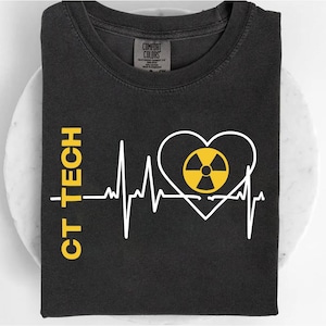 May include: Dark gray t-shirt with the text "CT TECH" in yellow on the left side. A white heartbeat line design includes a heart shape with a yellow radiation symbol in the center. The shirt is folded.