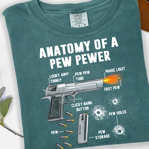 Könnte beinhalten: Türkisfarbenes T-Shirt mit dem Text "ANATOMY OF A PEW PEWER" und einer Abbildung einer Handfeuerwaffe mit Beschriftungen wie "LOOKY AIMY THINGY" und "MAGIC LIGHT". Das Design beinhaltet Patronenhülsen und Einschusslöcher.