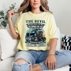 Puede incluir: Camiseta amarillo claro con un diseño gráfico. El diseño incluye un camión, la Parca y el texto "The Devil Whispered to Me 'I'm Coming For You' I Whisper Back Bring Beer". Los colores son negro, blanco y turquesa.