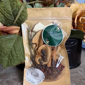 Könnte beinhalten: Ein durchsichtiger Kunststoffbeutel mit getrockneten Potpourri-Zutaten, darunter Zimtstangen, getrocknete Apfelscheiben, ganze Pimentbeeren und ein Rosmarinzweig. Der Beutel hat ein grünes, rundes Etikett mit der Aufschrift Kelli's Cozy Simmers und ein weißes Etikett mit dem Wort Fall.
