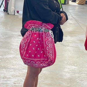 May include: A bright pink drawstring bag featuring a white paisley bandana pattern. The bag has a gathered top opening with a white cord closure and a decorative floral fabric trim along the inner edge of the drawstring channel.