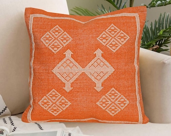 May include: Orange square throw pillow with a white geometric pattern. The pillow has a textured appearance and a white border. The design features diamond and arrow-like shapes. The pillow is placed on a white sofa.