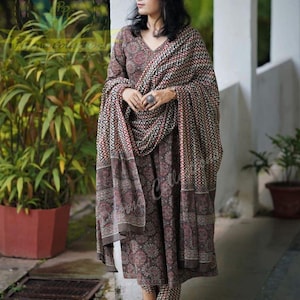 Puede incluir: Un conjunto salwar kameez estampado en marrón y rojo con un chal a juego. El conjunto incluye una parte superior con cuello en V, pantalones y un chal con un patrón geométrico. El atuendo se usa con zapatos marrones.