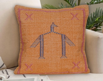 May include: Orange square throw pillow with a geometric design. The pillow features a central blue and orange figure, pink border, and pink X-shaped accents in each corner. The pillow is on a white sofa.