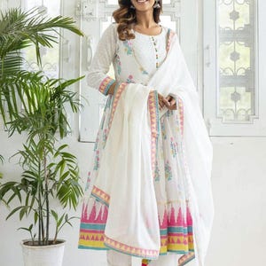 May include: White Indian-style suit with floral embroidery and colourful trim. The outfit includes a long-sleeved tunic, matching trousers, and a sheer scarf. The trim features pink, yellow, and blue accents. The outfit is worn with clear heels.