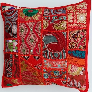 May include: A square, patchwork pillow with a red border. The pillow features a variety of red fabric squares, each with unique embroidered patterns in shades of red, green, blue, and gold. The seams are stitched with red thread.