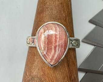 Puede incluir: Anillo de plata con una gran gema de rodocrosita en forma de lágrima. La piedra rosa y blanca está engastada en un bisel. El anillo tiene detalles decorativos en la banda.