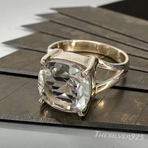 May include: A silver ring featuring a large, square, clear, faceted gemstone. The ring has a thick band and a setting that secures the gemstone. It is displayed on a textured, grey surface. The text "TheSilver925" is visible.