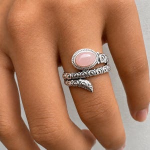 May include: A silver bypass ring featuring a smooth, oval-shaped light pink stone set in a decorative bezel. The band has a textured, coiled design with intricate scrollwork patterns that wrap around the finger in a spiral, open-ended style.