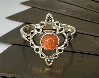 May include: A silver ring with an ornate, openwork design surrounding a round, orange-colored gemstone. The ring's band is smooth and polished. The gemstone has a speckled appearance. The ring is set against a dark background.