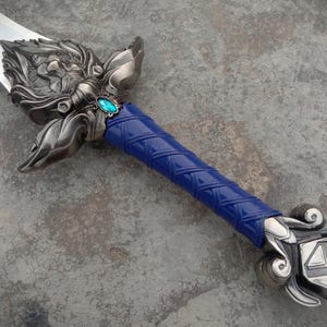 May include: A detailed fantasy sword with a silver blade and ornate metalwork. The hilt features a blue, textured grip and a decorative guard with a teal-colored gem. The sword's design is intricate and detailed.
