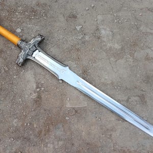May include: A silver and gold-toned sword with a light brown handle. The blade is long and sharp, and the hilt features ornate detailing. The sword is lying on a dirt surface.