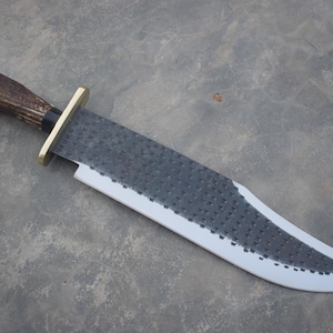Puede incluir: Un cuchillo grande, hecho a mano, con una hoja texturizada de color gris oscuro y un borde gris claro. El mango es de material de asta marrón y está asegurado con una guarda de latón. El cuchillo está diseñado para uso en exteriores.