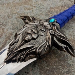 May include: A detailed, silver-colored sword with a lion's head design on the hilt. The handle is wrapped in blue material, and a blue gem is set near the blade. The blade is polished and reflective.