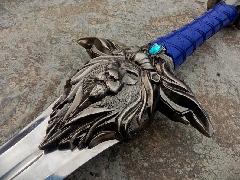May include: A detailed, silver-colored sword with a lion's head design on the hilt. The handle is wrapped in blue material, and a blue gem is set near the blade. The blade is polished and reflective.