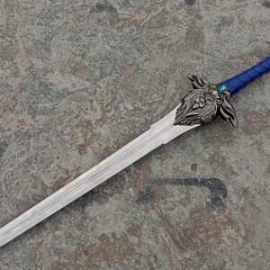 May include: A detailed fantasy sword with a silver blade and ornate hilt. The hilt features a blue wrapped grip, a decorative guard with floral and animal motifs, and a jewel. The sword is displayed on a textured gray surface.