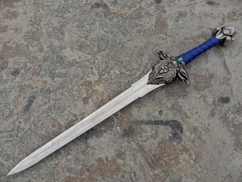 May include: A detailed fantasy sword with a silver blade and ornate hilt. The hilt features a blue wrapped grip, a decorative guard with floral and animal motifs, and a jewel. The sword is displayed on a textured gray surface.