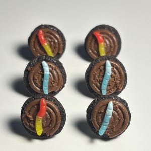 May include: Six chocolate cookies with brown frosting and gummy worms. The gummy worms are red, yellow, and blue. The cookies are arranged in two rows of three on a white surface.