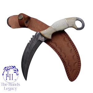 May include: A curved, Damascus steel knife with a bone handle and a brown leather sheath. The knife has a serrated edge and a ring at the end of the handle. The sheath is embossed with a repeating pattern. The Hands Legacy logo is in the lower left corner.