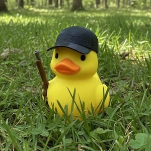 May include: A yellow rubber duck wearing a black baseball cap and holding a brown toy rifle, set in a grassy field with a forest background. The duck has an orange beak and black eyes.