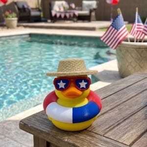 May include: A yellow rubber duck wearing a tan straw hat and red-framed sunglasses with white star lenses. The duck is positioned inside a red, white, and blue striped life preserver ring, featuring a bright orange beak and a smooth, rounded body.