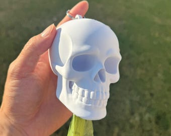 May include: A white skull-shaped dog waste bag dispenser with a silver keyring. The dispenser is held in a hand against a background of green grass and a wooden fence. A green bag is visible at the bottom.