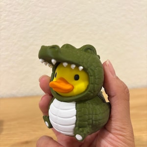 Puede incluir: Un pato de goma amarillo con un disfraz de cocodrilo verde texturizado, vientre blanco y dientes blancos. El pato tiene un pico naranja brillante y ojos negros, mientras que la capucha de cocodrilo presenta un patr&oacute;n de escamas y garras.