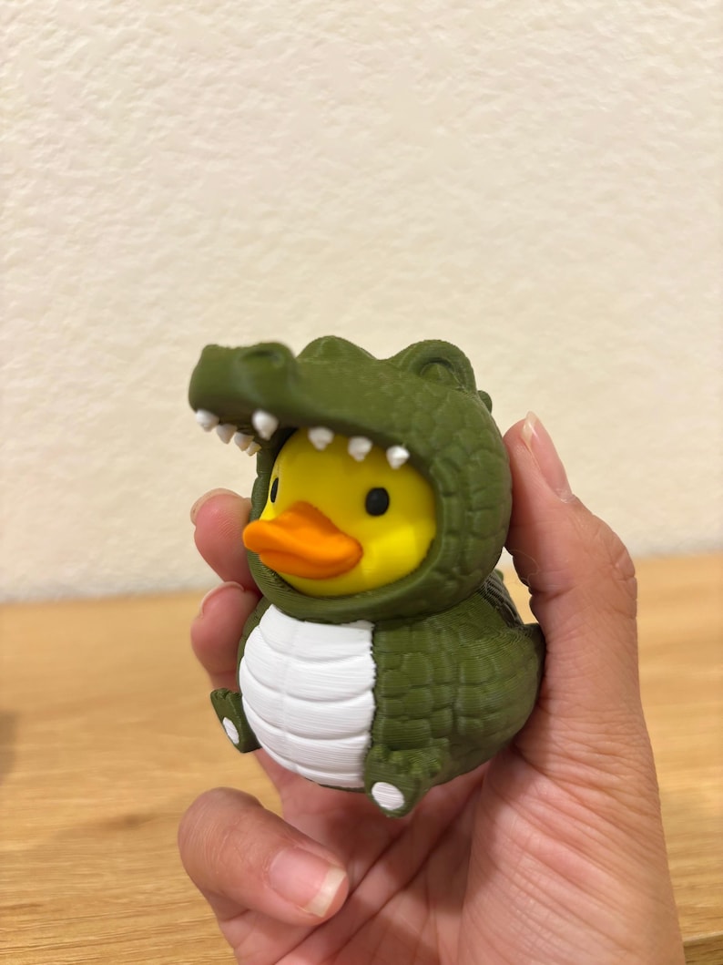 Puede incluir: Un pato de goma amarillo con un disfraz de cocodrilo verde texturizado, vientre blanco y dientes blancos. El pato tiene un pico naranja brillante y ojos negros, mientras que la capucha de cocodrilo presenta un patr&oacute;n de escamas y garras.