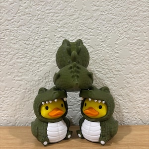 Puede incluir: Tres patos de goma amarillos con disfraces de caim&aacute;n verde oscuro, dientes blancos y escamas blancas en el vientre. Los dos patos inferiores est&aacute;n uno al lado del otro, mientras que el tercero est&aacute; encima, mostrando su espalda y cola.