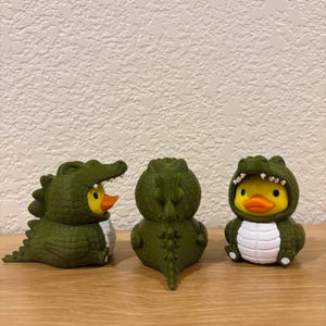 Puede incluir: Tres peque&ntilde;as figuras de pato de goma vestidas con disfraces de caim&aacute;n verde oscuro. Los patos tienen cuerpos amarillos y picos naranjas, mientras que los disfraces presentan escamas verdes texturizadas, vientres blancos y dientes blancos.