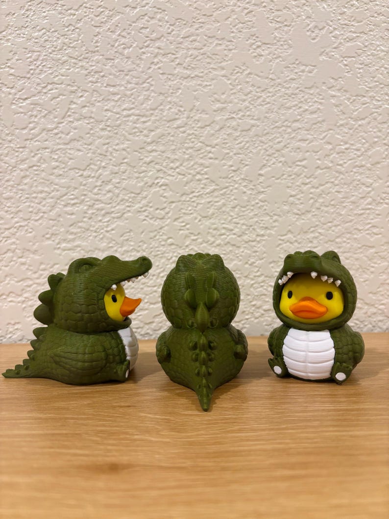 Puede incluir: Tres peque&ntilde;as figuras de pato de goma vestidas con disfraces de caim&aacute;n verde oscuro. Los patos tienen cuerpos amarillos y picos naranjas, mientras que los disfraces presentan escamas verdes texturizadas, vientres blancos y dientes blancos.