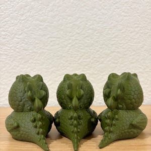 Puede incluir: Tres peque&ntilde;as figuras de dinosaurios de color verde oliva dispuestas en fila. Cada figura presenta una superficie texturizada con escamas, una cabeza redondeada y una cresta prominente de espinas a lo largo del lomo hasta la cola.