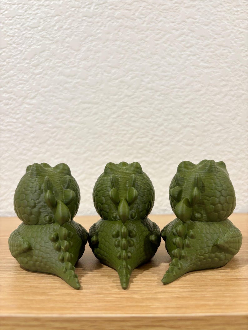 Puede incluir: Tres peque&ntilde;as figuras de dinosaurios de color verde oliva dispuestas en fila. Cada figura presenta una superficie texturizada con escamas, una cabeza redondeada y una cresta prominente de espinas a lo largo del lomo hasta la cola.