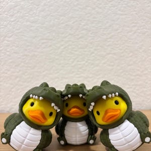 Puede incluir: Tres peque&ntilde;as figuras de patos de goma, cada una con un disfraz de caim&aacute;n verde oliva oscuro con vientre blanco y una capucha con dientes blancos. Los patos tienen caras de color amarillo brillante y picos naranjas, agrupados juntos.