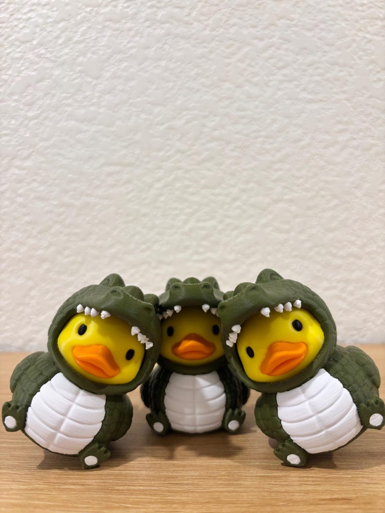 Puede incluir: Tres peque&ntilde;as figuras de patos de goma, cada una con un disfraz de caim&aacute;n verde oliva oscuro con vientre blanco y una capucha con dientes blancos. Los patos tienen caras de color amarillo brillante y picos naranjas, agrupados juntos.